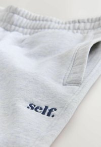 Light gray sweatpants with an elastic waistband and a small dark blue embroidered text "self." near the pocket.