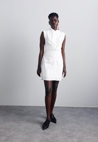 Vestito camicia bianco senza maniche con dettaglio annodato in vita, chiusura con bottoni sul davanti e vestibilità rilassata, abbinato a scarpe nere con texture.