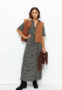 Un gilet en daim marron superposé sur une robe maxi à motifs paisley noirs. Des bottines noires et un sac à franges marron complètent le look.
