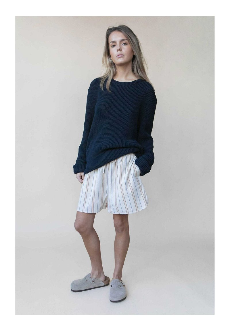 Navy gestrickter Pullover mit gerippter Textur, kombiniert mit weißen Streifen-Shorts und grauen Slippern, die ein lockeres, lässiges Design aufweisen.