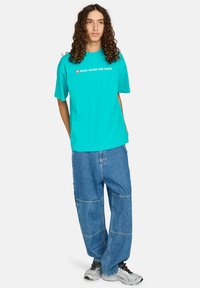 Young person with long curly hair wearing turquoise "WIND WATER FIRE EARTH" t-shirt, baggy patched blue jeans, and silver sneakers, standing.