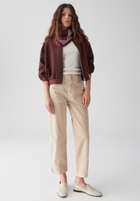 Eine Frau trägt eine bordeauxrote Zip-Jacke, ein weißes Oberteil, beigefarbene hoch taillierte Hose, beigefarbene Slipper und einen lila gemusterten Halstuch und steht vor einem schlichten Hintergrund.