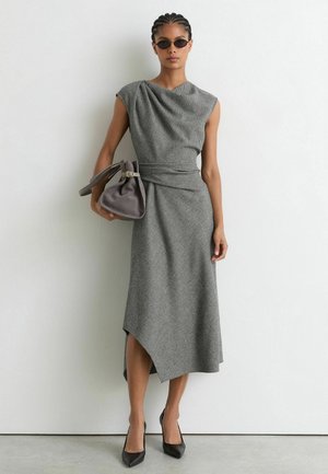 Frau trägt ein graues ärmelloses asymmetrisches Kleid, schwarze High Heels, Sonnenbrille und hält eine graue Handtasche vor einer weißen Wand.