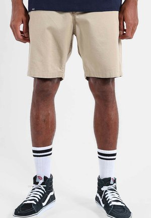 Jambes d'homme portant un short beige, des chaussettes blanches montantes avec des rayures noires, et des baskets montantes noires avec des lacets blancs.