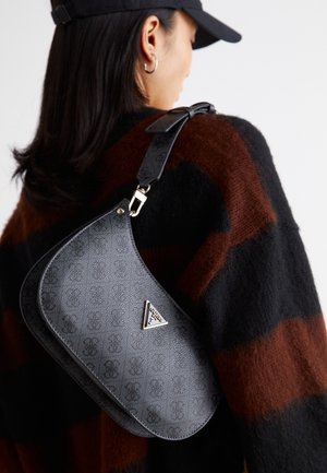 Personne portant un pull rayé noir et marron et une casquette noire, portant un sac à bandoulière en cuir noir avec un logo métallique triangulaire.