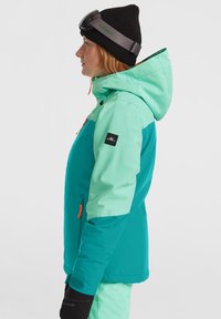 Teal en mint groene split snowboardjas met een ritszak, oranje accenten, capuchon en zwarte handschoenen. Draagt een zwarte beanie en skibril.