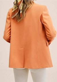 Blazer orange cintré avec épaules structurées, texture lisse et fente dans le dos. Stylé avec une écharpe à motifs nouée au cou.