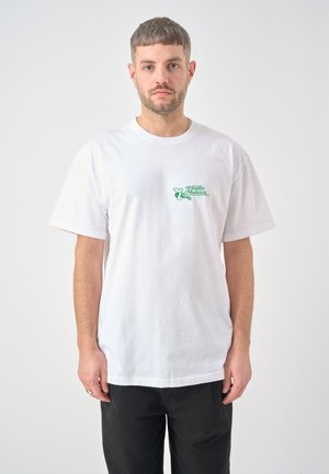 Man met kort bruin haar en baard, draagt een wit T-shirt met een groen logo op de borst en zwarte broek, staand tegen een effen witte achtergrond.