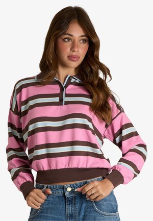 Donna che indossa un maglione a righe rosa, marrone e blu a maniche lunghe con collo e bottoni, abbinato a jeans blu.