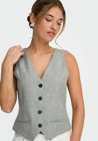 Jeune femme aux cheveux châtain clair coiffés en chignon, portant un gilet gris sans manches boutonné et un pantalon blanc, accessoirisée avec des boucles d'oreilles en forme de fleur et une bague.