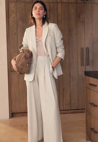 Sivo odijelo s finim prugama, široke hlače i uska blejzerica, upareni s čipkanim topom i smeđom torbom od brušene kože. Drvena pozadina.