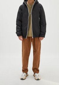 Veste matelassée noire avec capuche et fermeture éclair blanche, associée à un pantalon en velours côtelé marron. Chaussures : baskets blanches et grises.