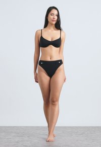 Μαύρο μπικίνι σετ με κορυφαίο τοπ cropped με λεπτές τιράντες και εσώρουχα χαμηλής μέσης με ασημένιες λεπτομέρειες, εμφανίζεται σε ανοιχτό φόντο.