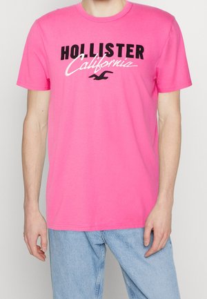 Pink cotton t-shirt with short sleeves. Features black lettering "HOLLISTER" and "California" in a cursive font. Smooth texture.