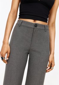 Pantalon gris sur mesure avec fermeture à bouton, passants de ceinture et tissu texturé. Des mains tenant une bague sont partiellement visibles.