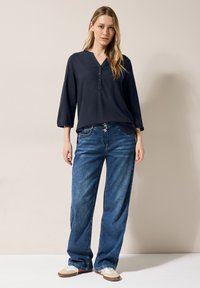 Dunkelblaue, langärmelige Bluse mit V-Ausschnitt und Knopfleiste, kombiniert mit hellblauen, weiten Jeans. Legere Schuhe runden das Outfit ab.