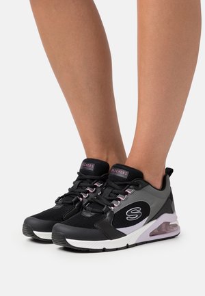 Des jambes portant des sneakers Skechers noirs et gris avec des accents roses et des semelles blanches, se tenant sur un fond blanc uni.