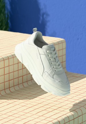 Witte leren sneaker met een chunky zool, voorzien van vetersluiting en trekband. Geplaatst tegen een rasterpatroon en een blauwe achtergrond.