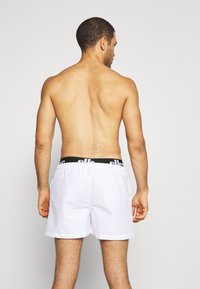 Weiße Schwimmshorts mit einem elastischen Bund, auf dem ein schwarzer Logo-Text zu sehen ist, von hinten auf einem nackten Oberkörper präsentiert. Glatte Oberfläche.