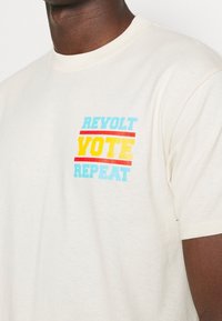 Krémová bavlněná tričko s grafickým potiskem v modré, žluté a červené barvě s textem "REVOLT VOTE REPEAT." Krátké rukávy a kulatý výstřih.