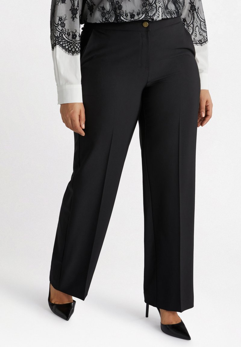 Svarta vida byxor i mjukt tyg, med framknäppning och skarpa veck, tillsammans med svarta spetsiga klackar.