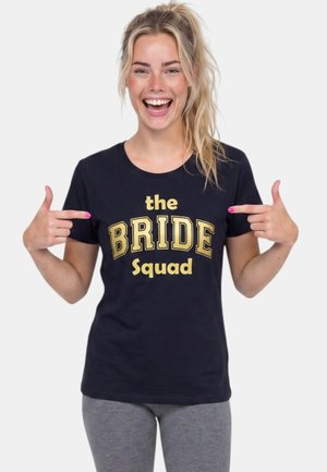 Mujer sonriente con cabello rubio recogido en una coleta señala su camiseta negra con la inscripción "the BRIDE Squad" en letras doradas, vistiendo pantalones grises.