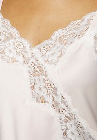 Rožinė satino camisole su sudėtinga balta nėriniu palei iškirptę ir pečius, minkšta, lygi tekstūra ir subtiliais gėlių raštais.