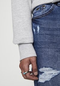 Camisa de manga larga gris y jeans azules desgastados. La mano lleva un anillo de plata con una piedra turquesa; los jeans tienen acentos rasgados cerca de la rodilla.