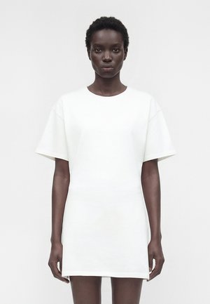 Femme noire mince aux cheveux courts bouclés, portant une robe t-shirt blanche unie et ample, debout devant un fond blanc.