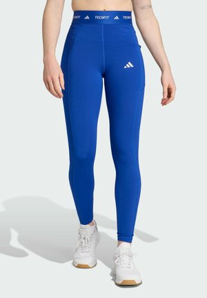 Plave Techfit tajice s visokim pojasom, izrađene od rastezljivog materijala. Sadrže bijeli Adidas logo na pojasu i bedrima.