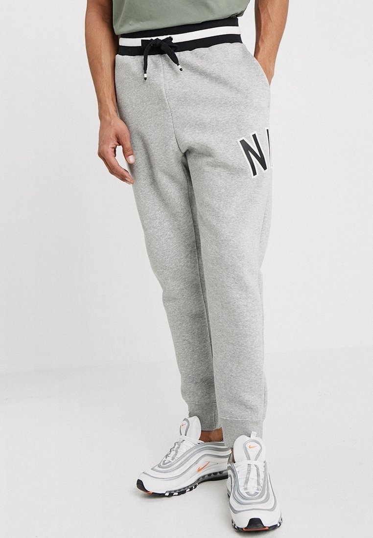 Šedé tepláky s černo-bílým pruhovaným pasem, na levém stehně s velkým logem "N". V páru s bílými sportovními botami.