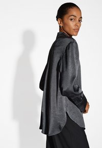 Camicia nera a maniche lunghe con texture, caratterizzata da un orlo curvo. Presenta una finitura liscia e lucida e un colletto.