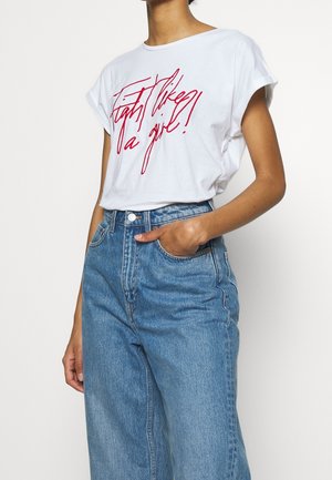 Biały t-shirt z podwiniętymi rękawami i czerwonym napisem "Walka jak dziewczyna!" noszony z niebieskimi dżinsami z wysokim stanem, ręka w kieszeni, brak widocznej głowy.