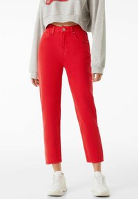 Pantalones vaqueros rojos de cintura alta, con un corte recto, cinco bolsillos y un acabado limpio. Combinados con zapatillas blancas y un suéter corto gris.
