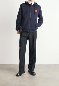 Hombre con sudadera azul marino con una etiqueta roja "Hugo", camisa blanca, pantalones oscuros, zapatos negros, sosteniendo una cartera negra en una mano.