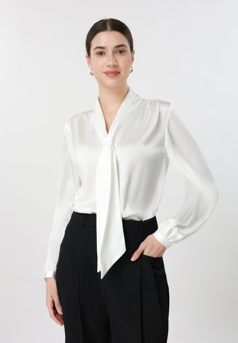 Femme aux cheveux foncés portant un chemisier en soie blanche avec un nœud au col, un pantalon noir taille haute et des boucles d'oreilles créoles dorées, debout, main dans la poche.