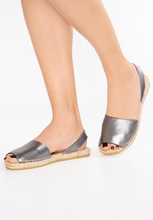 Stopy w sandałach typu slingback z otwartymi palcami w metalicznym srebrnym kolorze, z plecionymi podeszwami ze sznurka jutowego, na białym tle.