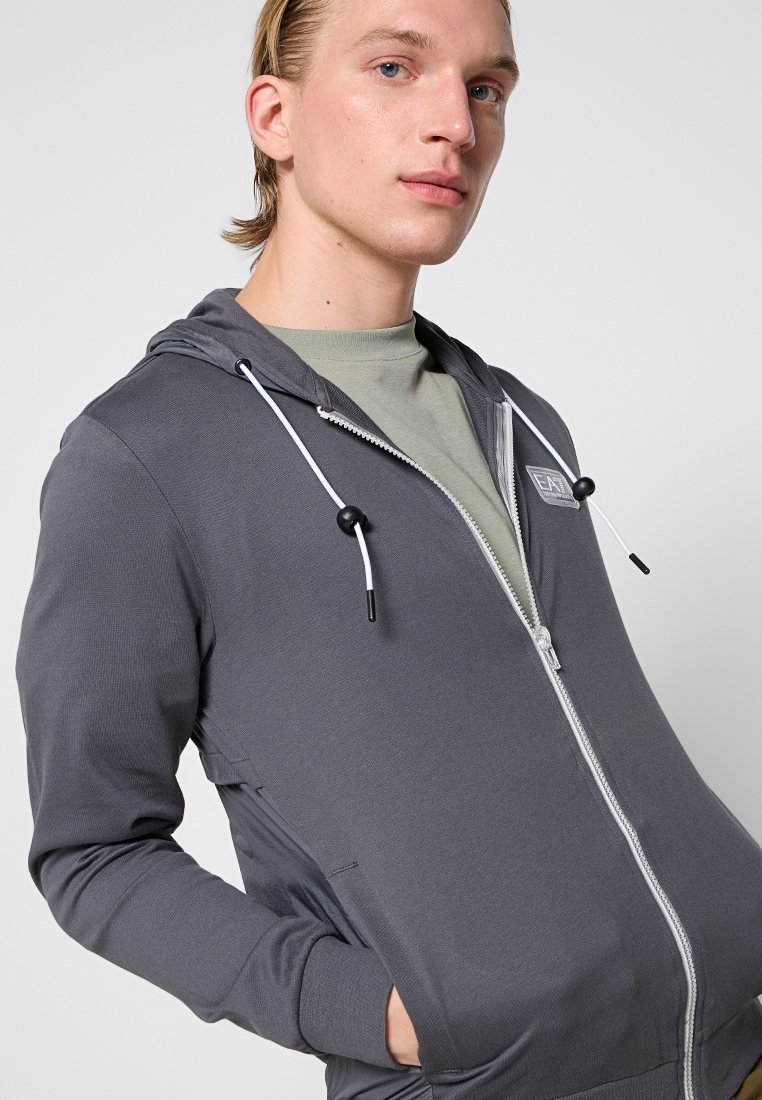 Felpa grigia con zip e cordino, tessuto texture, due tasche laterali e un patch con logo. Indossata sopra una maglietta verde chiaro.