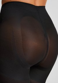 Nahaufnahme einer Person, die schwarze transparente Shaping-Tights trägt, die den verstärkten Gesäßbereich und die Taillennaht betonen.