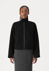 Black fuzzy bomber jacket with a front zipper, round neckline, and loose-fit sleeves, paired with a textured gray ribbed skirt.