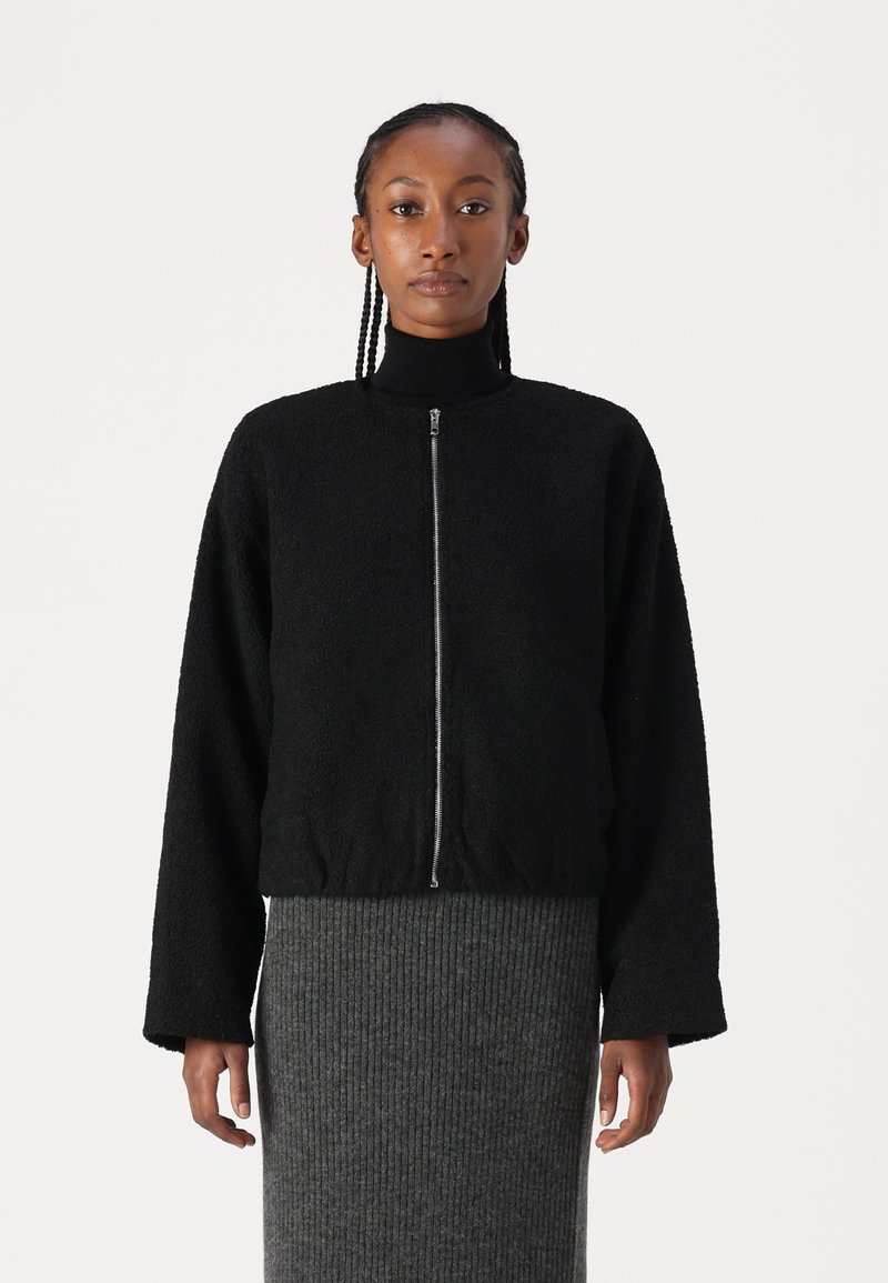 Black fuzzy bomber jacket with a front zipper, round neckline, and loose-fit sleeves, paired with a textured gray ribbed skirt.