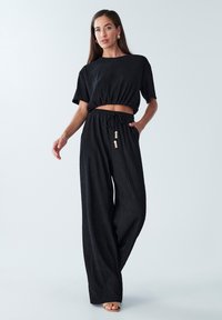 Černý žebrovaný crop top s krátkými rukávy, spárovaný s širokými kalhotami. Nastavitelný pas s vázáním a korálkovými akcenty. Hladká textura.