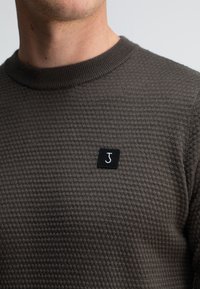 Nahaufnahme einer Person, die einen strukturierten dunkelgrauen Pullover mit einem schwarzen Patch trägt, auf dem ein weißer Buchstabe "J" auf der Brust zu sehen ist.