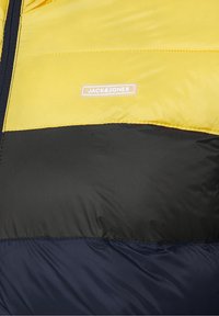 Sección de chaqueta acolchada en amarillo, negro y azul marino con costuras horizontales y una etiqueta blanca con el logo "JACK & JONES" en el panel amarillo.