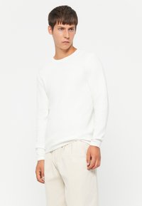 Witte, gestructureerde blouse met lange mouwen en een ronde hals, gedragen met lichte pantalons. De stof heeft een subtiel patroon en een relaxte pasvorm.