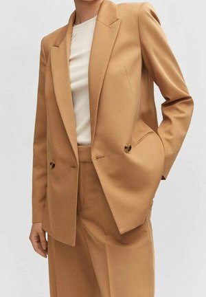 Femme portant un costume beige sur mesure avec un blazer à double bouton et un pantalon taille haute assorti, porté sur un haut blanc à col rond.