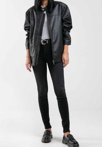 Chaqueta de piel sintética negra con cremallera frontal, corte relajado y hombros estructurados, combinada con vaqueros ajustados negros y zapatos negros sin cordones.