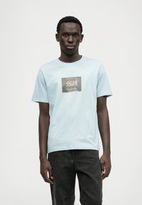 Svetlomodré bavlnené tričko s námestným grafickým potlačou, na ktorej je nápis "JACK & JONES" v bielej a oranžovej farbe. Krátke rukávy a okrúhly výstrih.