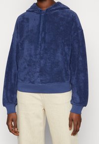 Granatowy polarowy sweatshirt z kapturem, z sznurkiem do regulacji, ściągaczami przy mankietach i na dole, zaprezentowany na tle jasnych spodni.