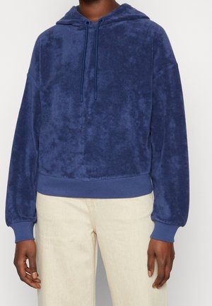 Felpa con cappuccio blu navy in tessuto soffice, con coulisse, polsini a costine e orlo, indossata sopra pantaloni chiari.
